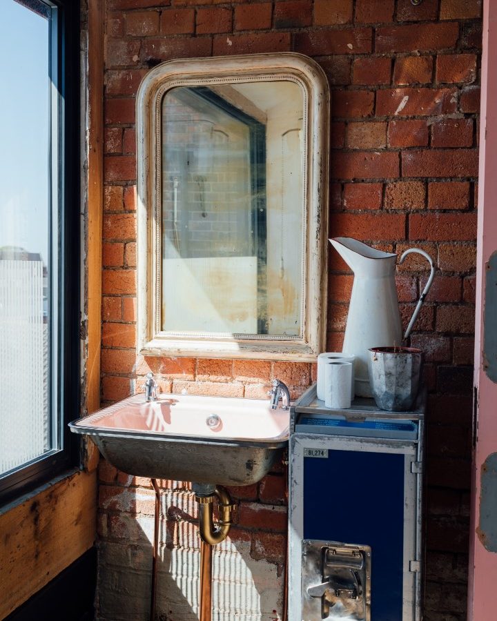 Vintage sink with a mirror on a brick wall, a white pitcher, and a British Airways metal cart nearby, in a sunlit room.