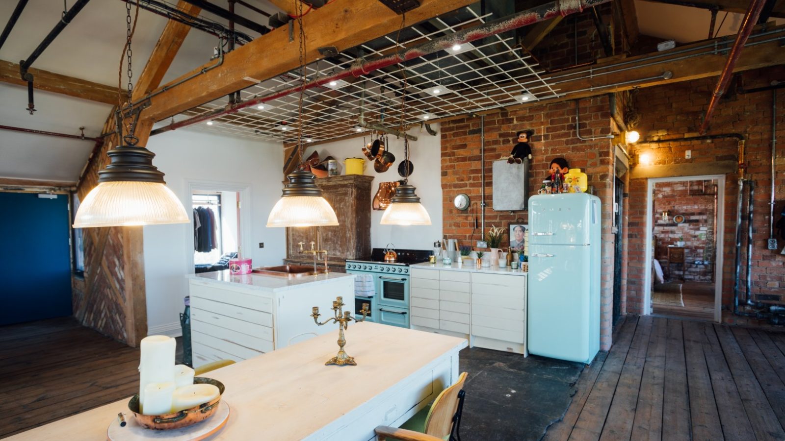 Open-concept kitchen with exposed beams, vintage appliances, brick walls, and industrial lighting over a white dining table.