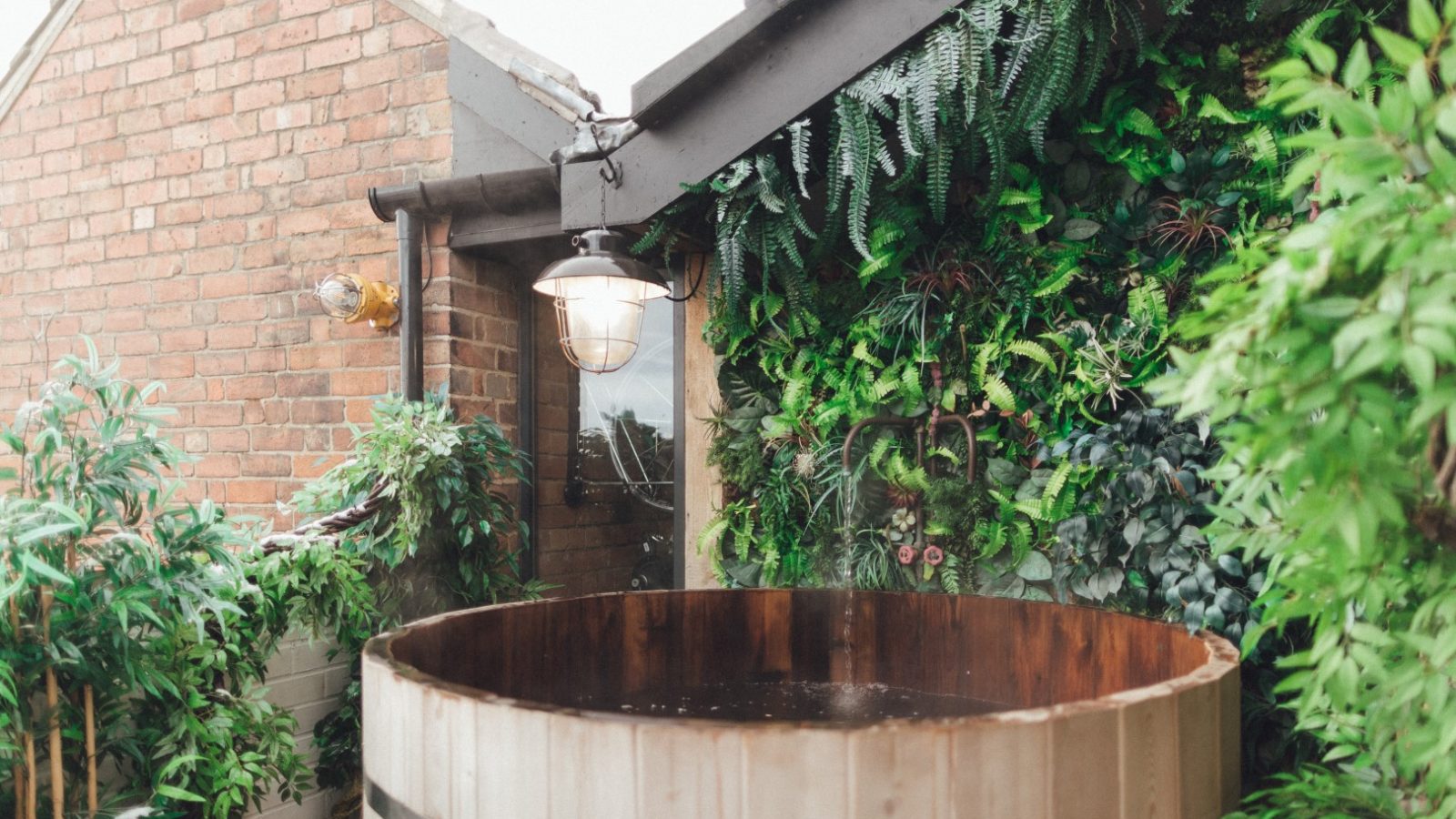 Wooden hot tub surrounded by lush greenery and brick walls, with overhead lighting.