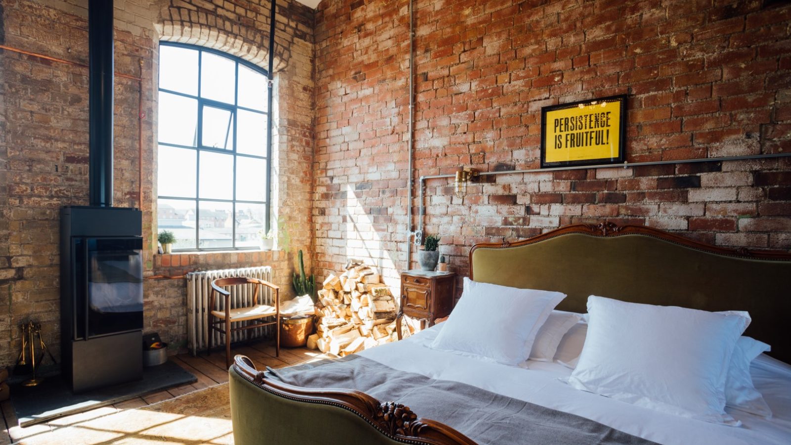 Loft-style bedroom with exposed brick, large window, fireplace, green bed, and motivational sign on the wall.