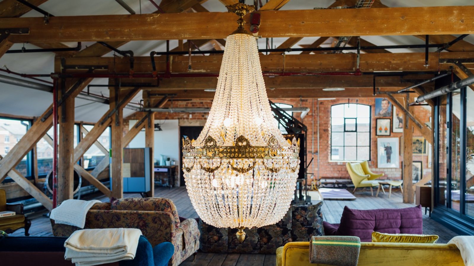 Spacious loft with a large crystal chandelier, exposed beams, colorful furniture, wooden floor, and brick walls.