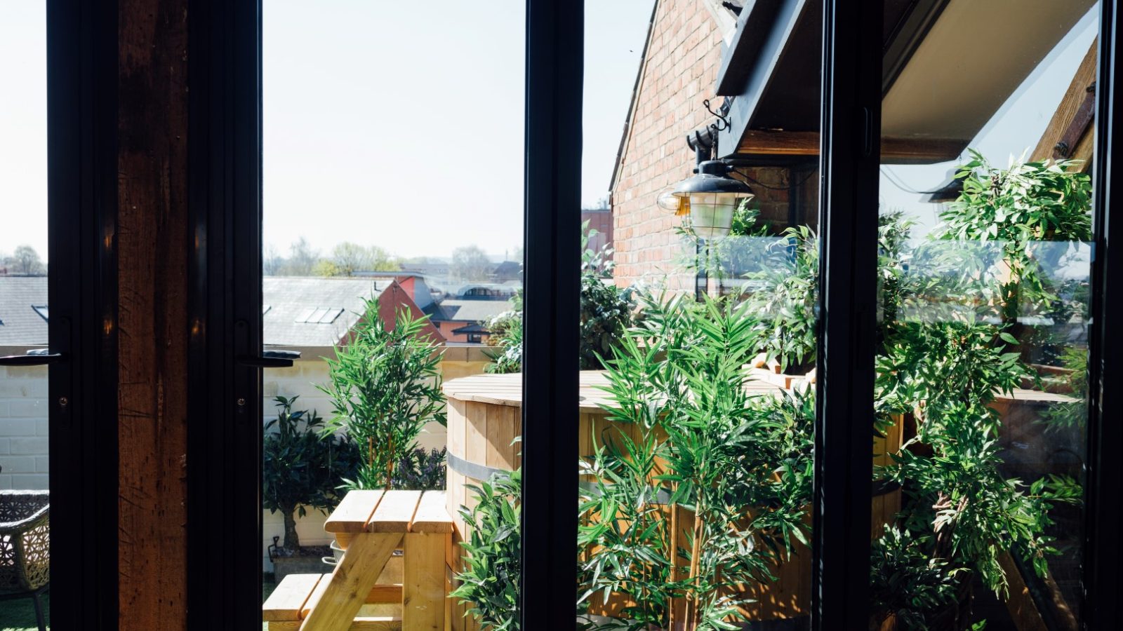 View through glass doors of a rooftop patio with plants, a wooden picnic table, and distant rooftops under a clear sky.