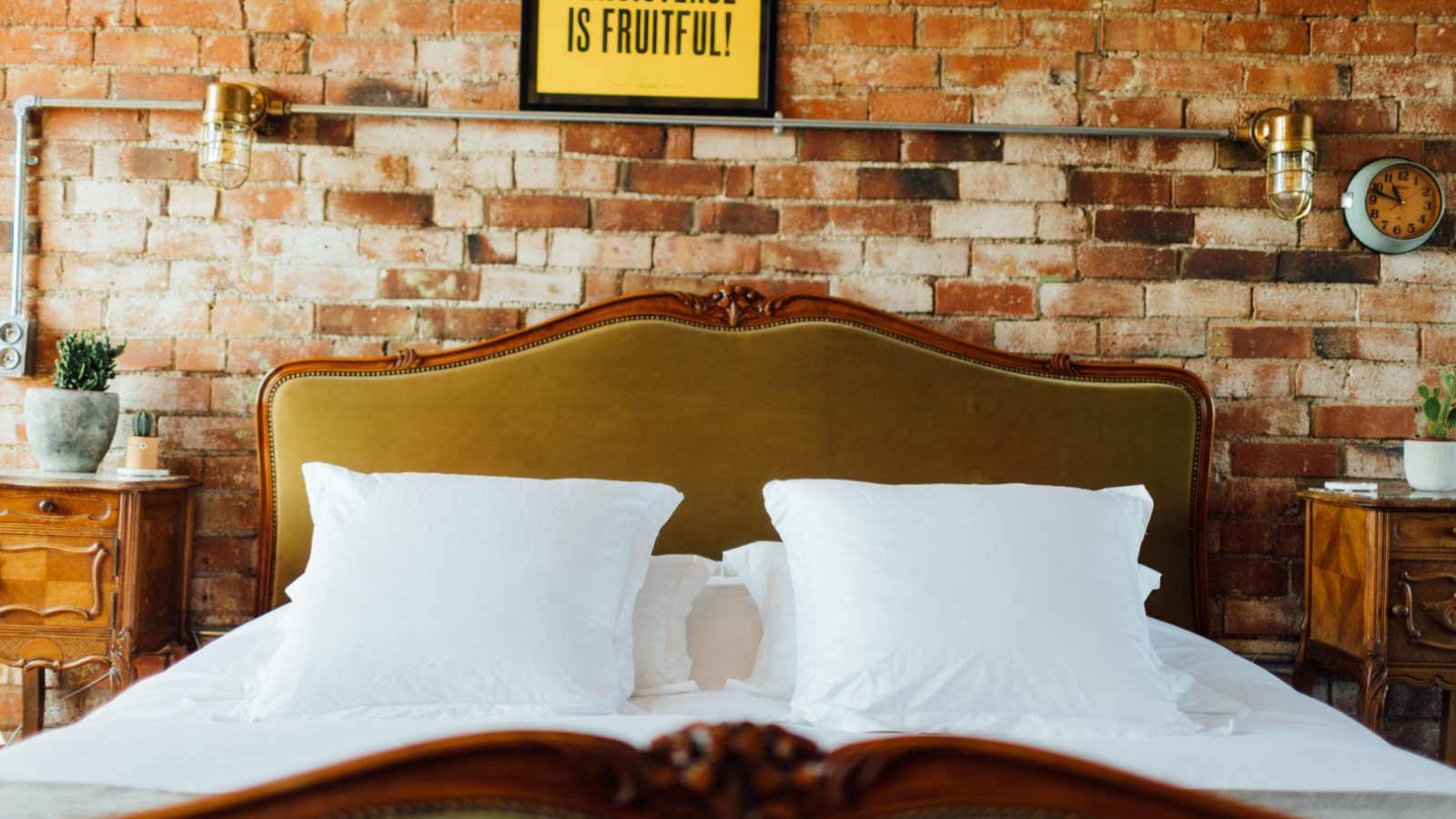 A bed with white pillows is set against a brick wall. Above, a sign reads 