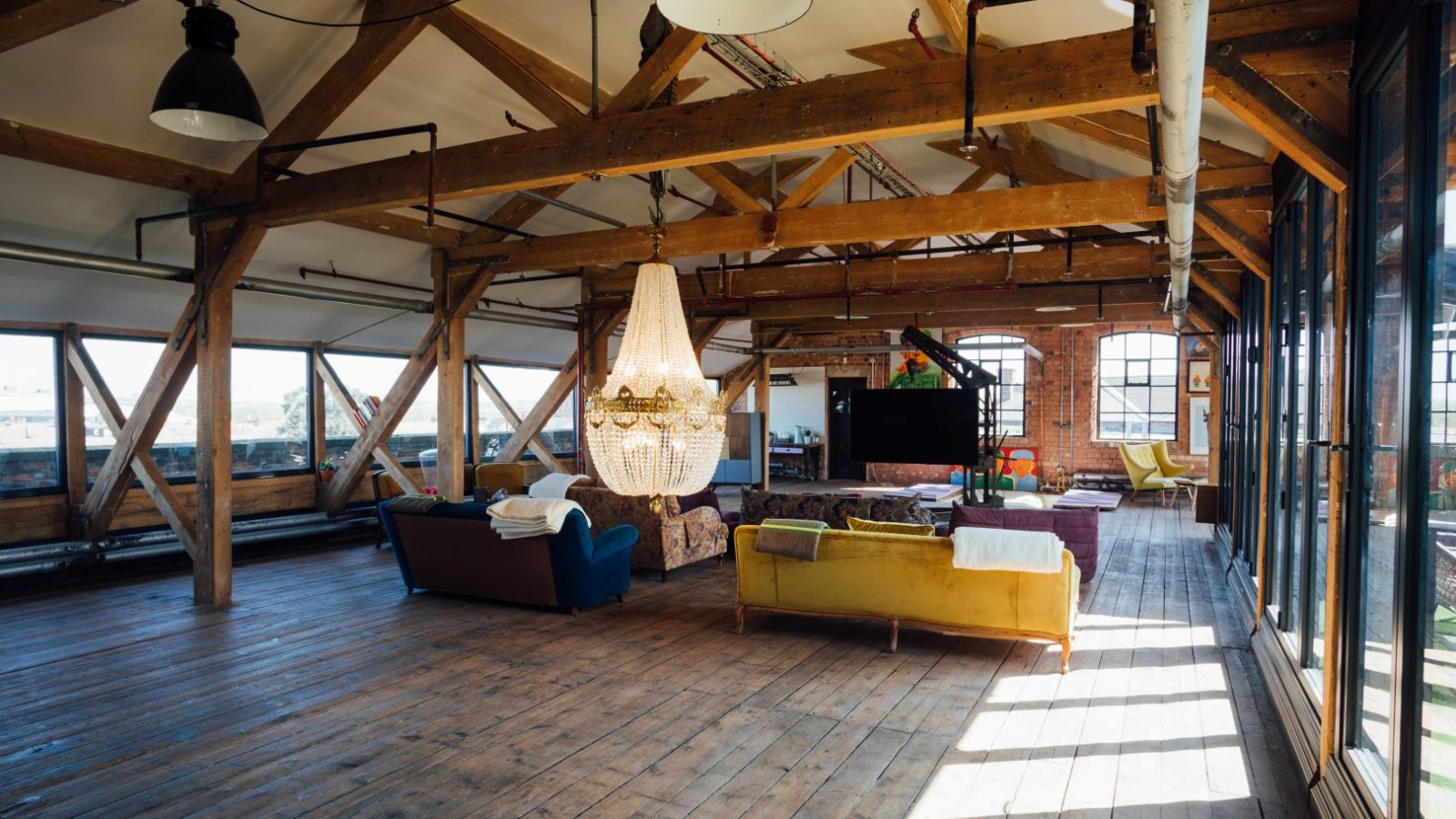 Spacious loft with exposed wooden beams, large windows, mixed seating, and a chandelier, featuring a wooden floor.