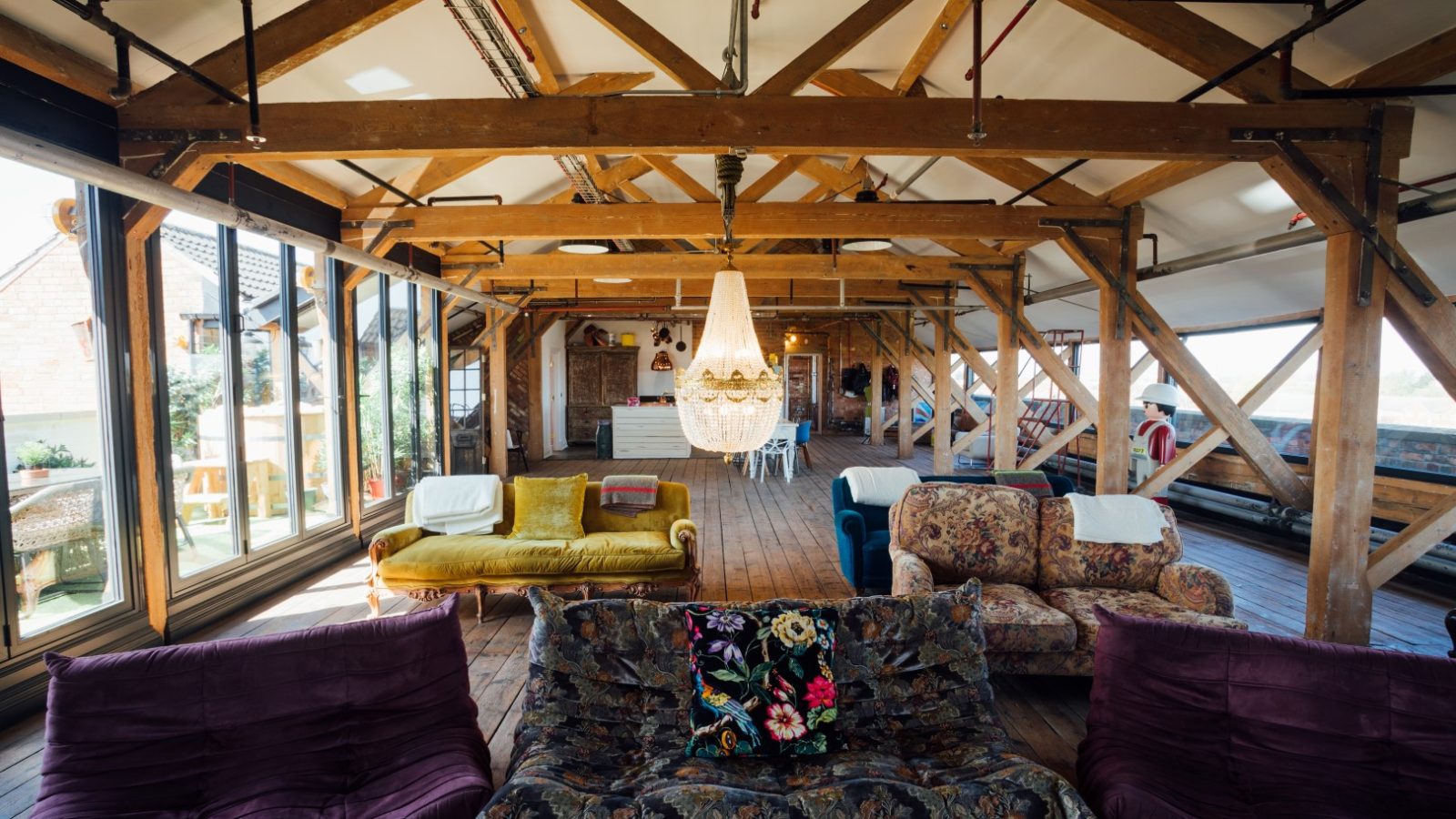 Spacious loft with wooden beams, eclectic sofas, a large chandelier, and natural light from large windows.