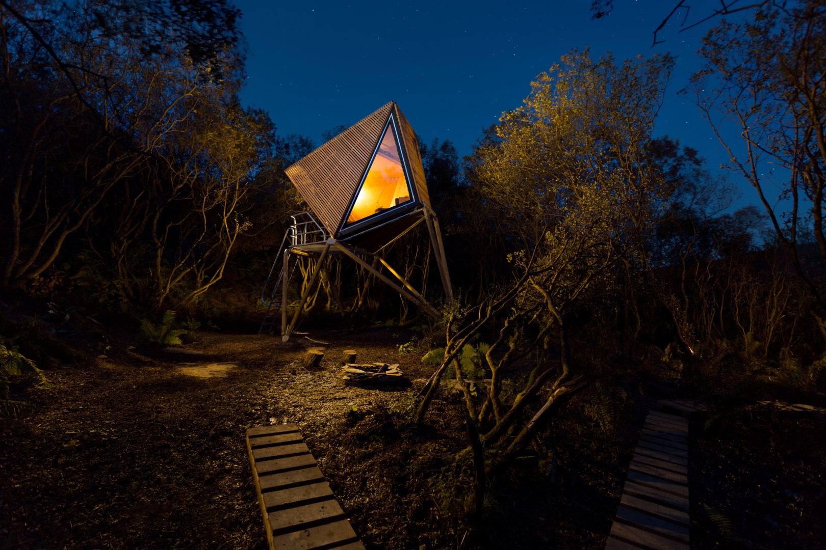 Digital detox break at this off grid retreat. Elevated triangular cabin with large window, glowing amidst trees in a nighttime forest setting.