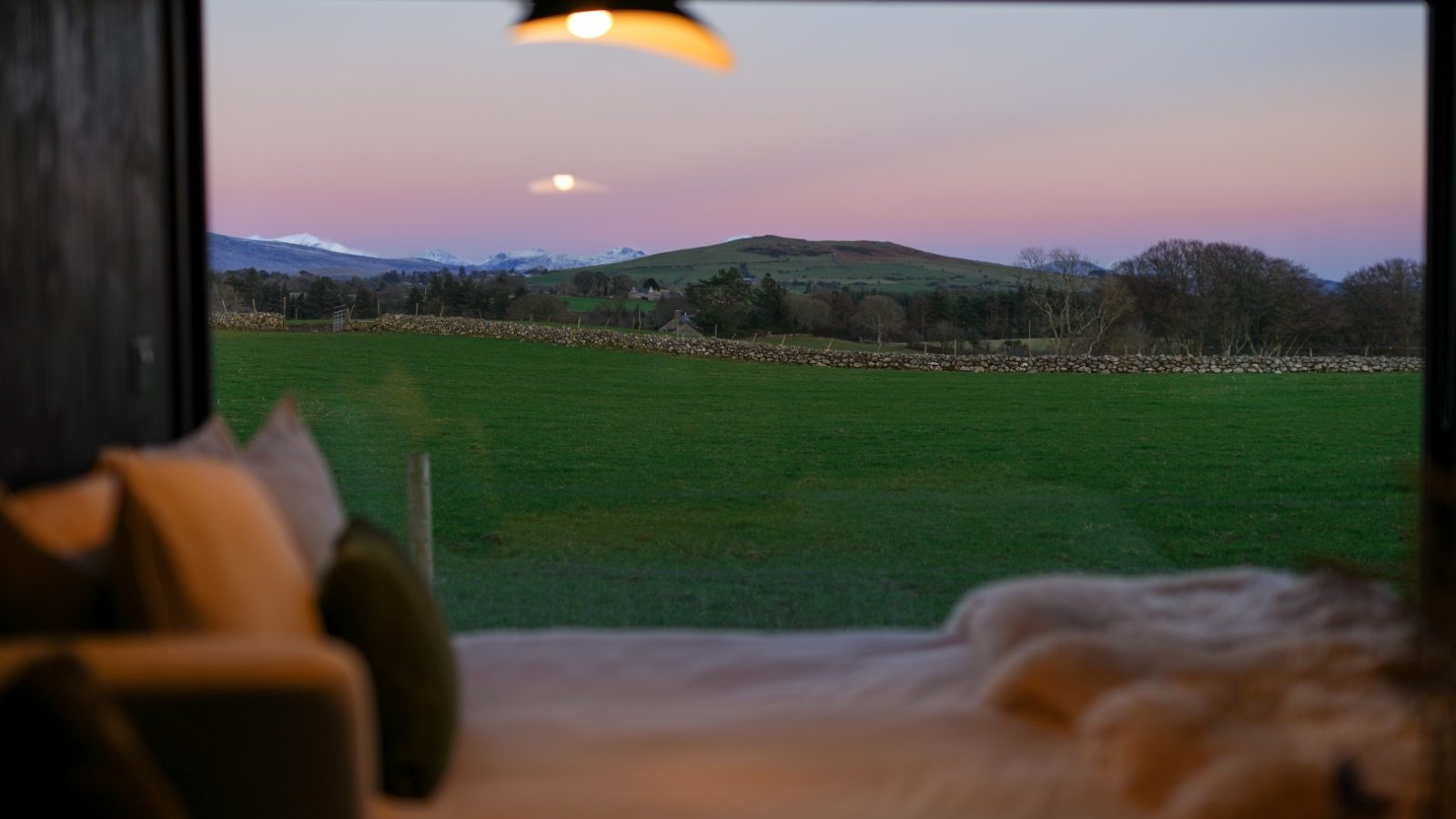 View from a window showing a peaceful landscape with green fields, distant mountains, and a colorful sunset sky.