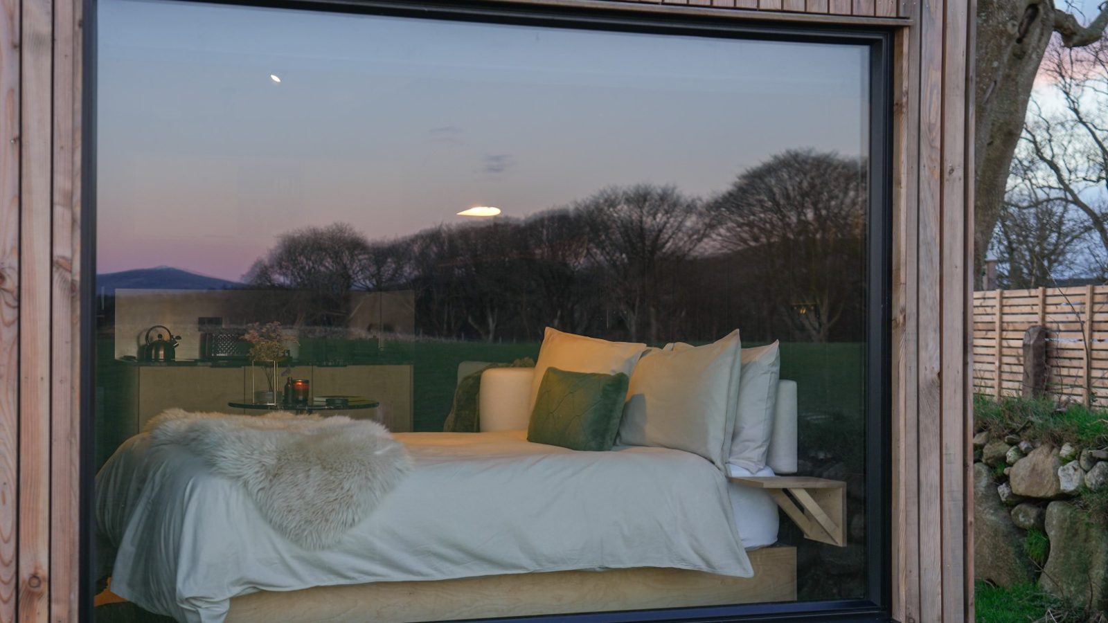 A cozy bedroom with a large window overlooks a scenic view of trees at dusk; the bed is adorned with pillows and a throw.