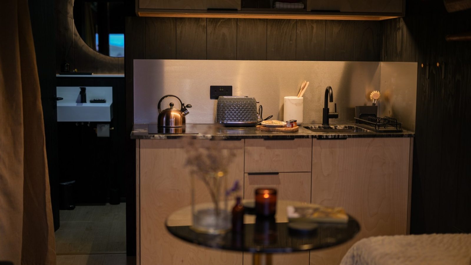 Cozy kitchenette with wooden cabinets, kettle, and toaster. A sink is underneath wooden shelves. A small round table is in front.