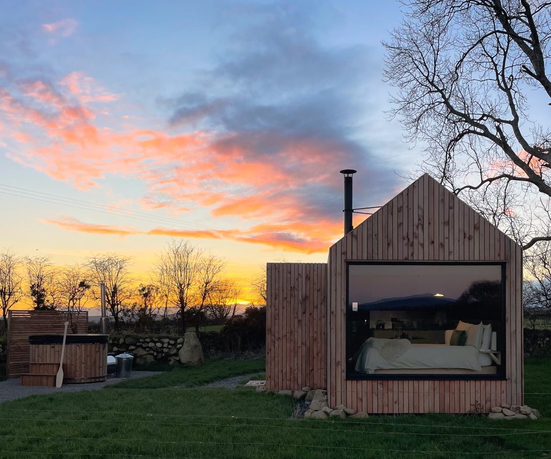 A wooden cabin with large windows stands in a grassy field at sunset, with a small hot tub nearby and trees in the background.