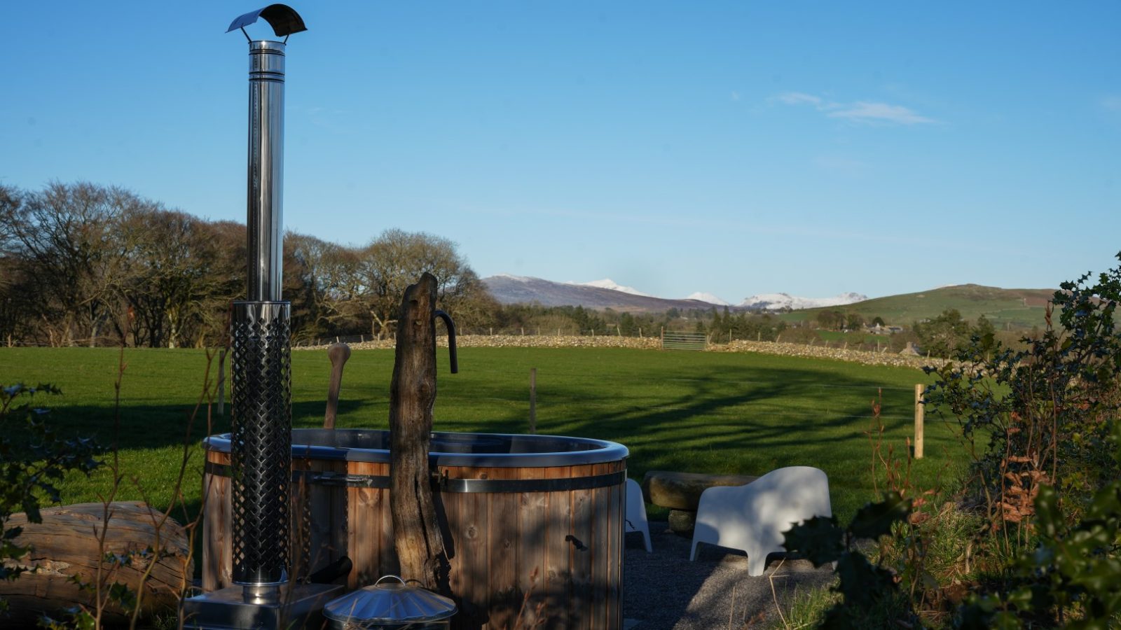 Wooden hot tub with tall chimney in a grassy field, surrounded by trees. Hills and a clear blue sky are in the background.