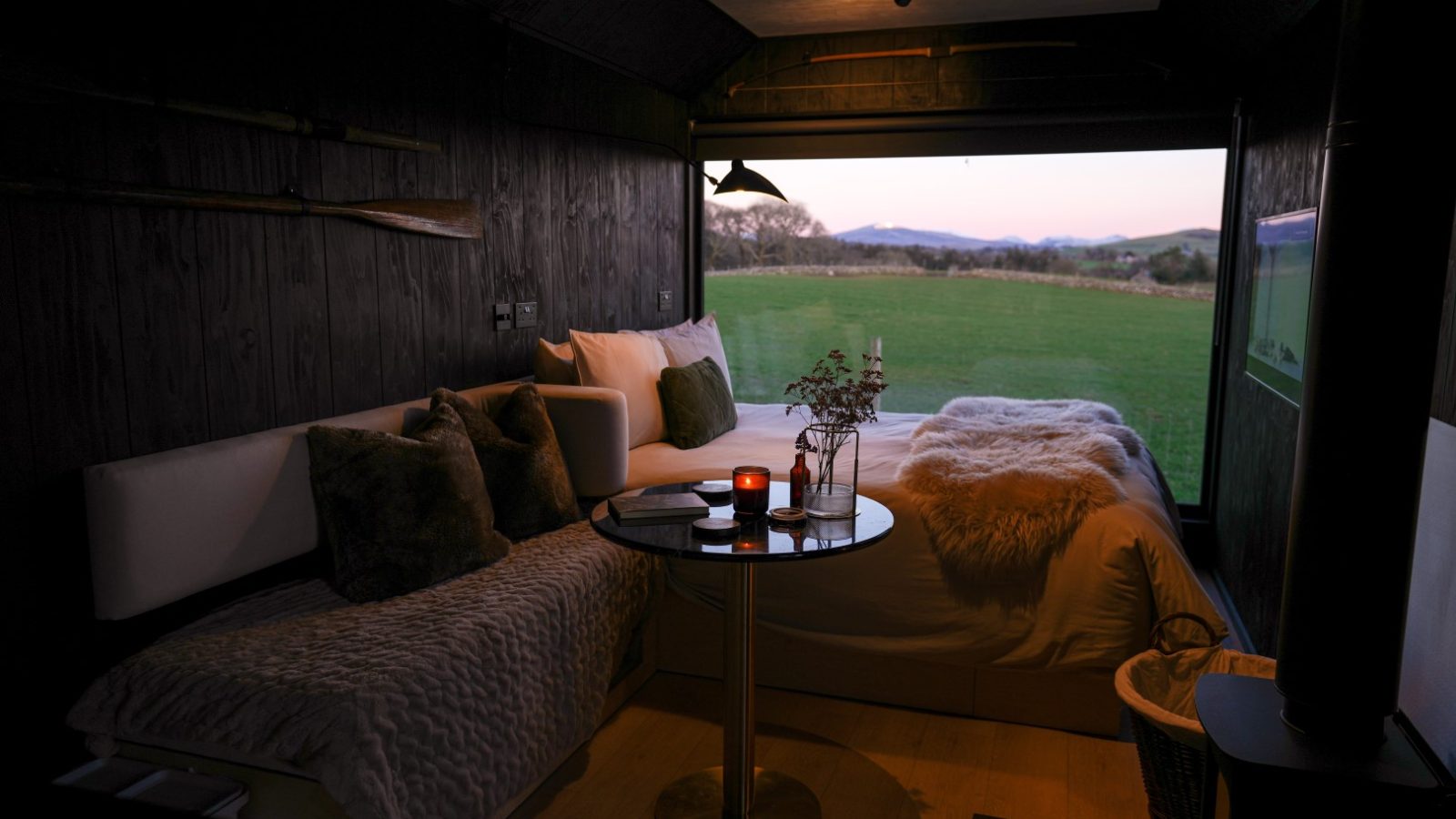 Cozy room with a sofa, bed, small table, and a large window revealing a scenic view of a grassy field and mountains at dusk.