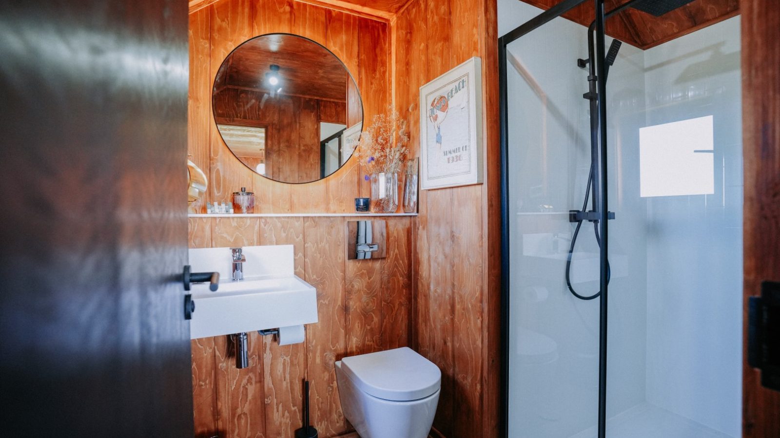 Small wooden bathroom with a round mirror, white sink, toilet, glass shower, and framed picture.