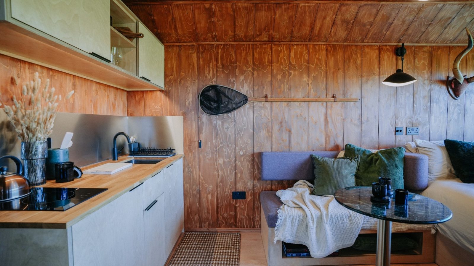 Compact wooden interior with a kitchenette, seating area, fishing net on the wall, and a small dining table with a camera.