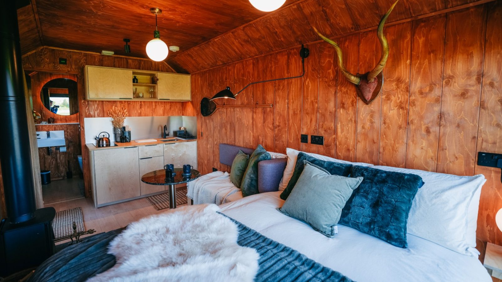 Cozy cabin interior with wooden walls, a bed, armchair, small kitchen, antler decor, and soft lighting.