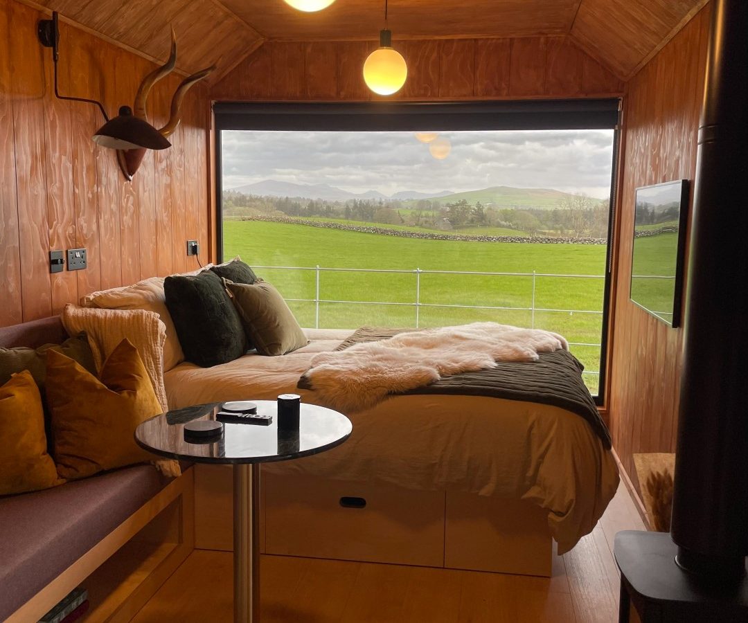 Cozy cabin interior with a bed, table, and large window view of a green field and cloudy sky. Warm lighting and wood decor.