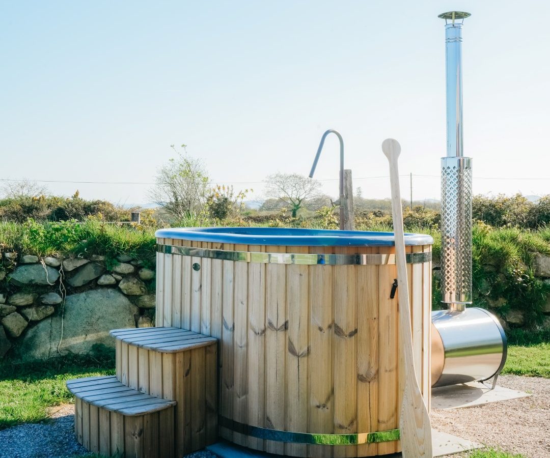 Wooden hot tub outdoors with a metal chimney, steps for access, and a wooden paddle on the side. Grass and stone wall nearby.