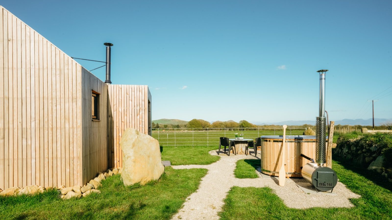 Wooden cabin with a chimney, hot tub, and patio seating on a grassy area. Open, rural landscape in the background under blue sky.