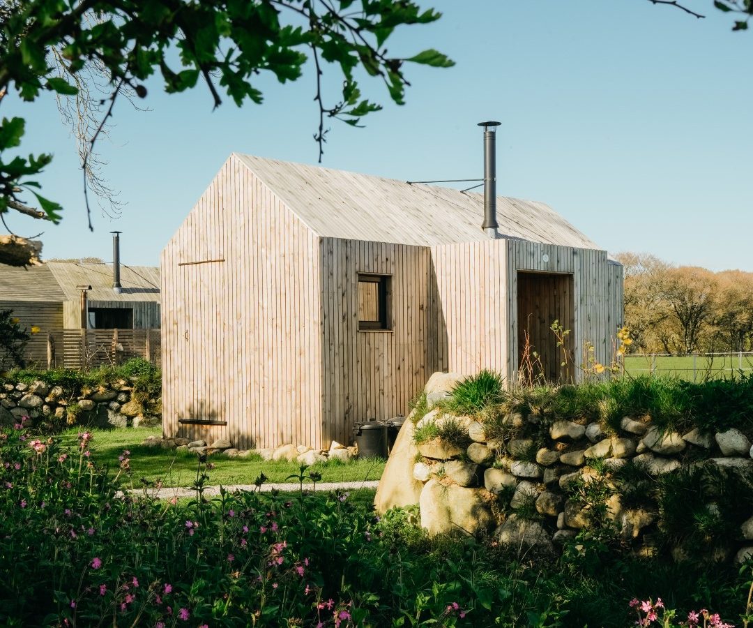 A small wooden cabin with a chimney stands in a garden surrounded by rocks and plants under a clear blue sky.