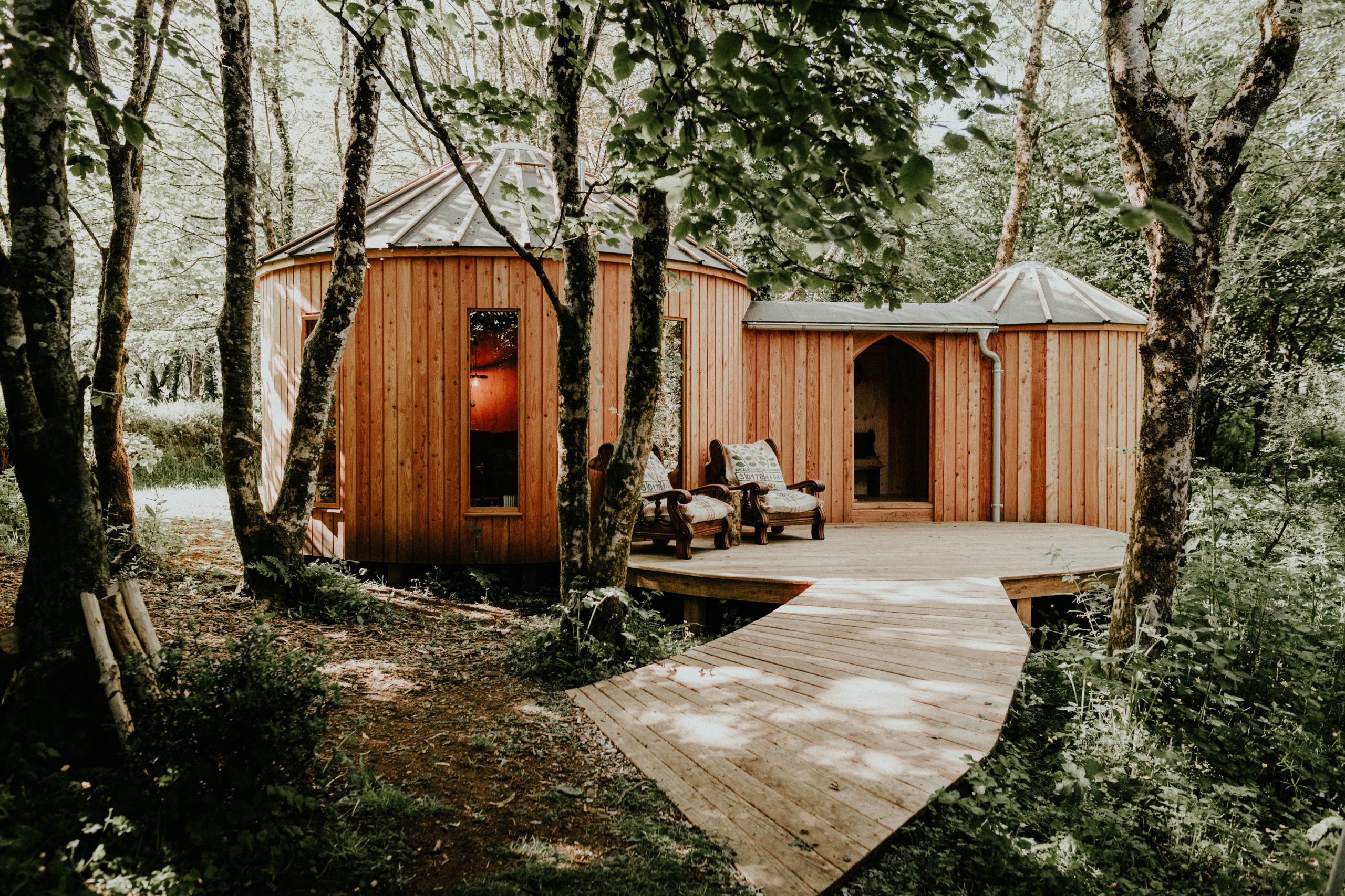 Wooden cabin with circular sections for digital detox stays, surrounded by trees and cozy chairs along a wooden walkway.