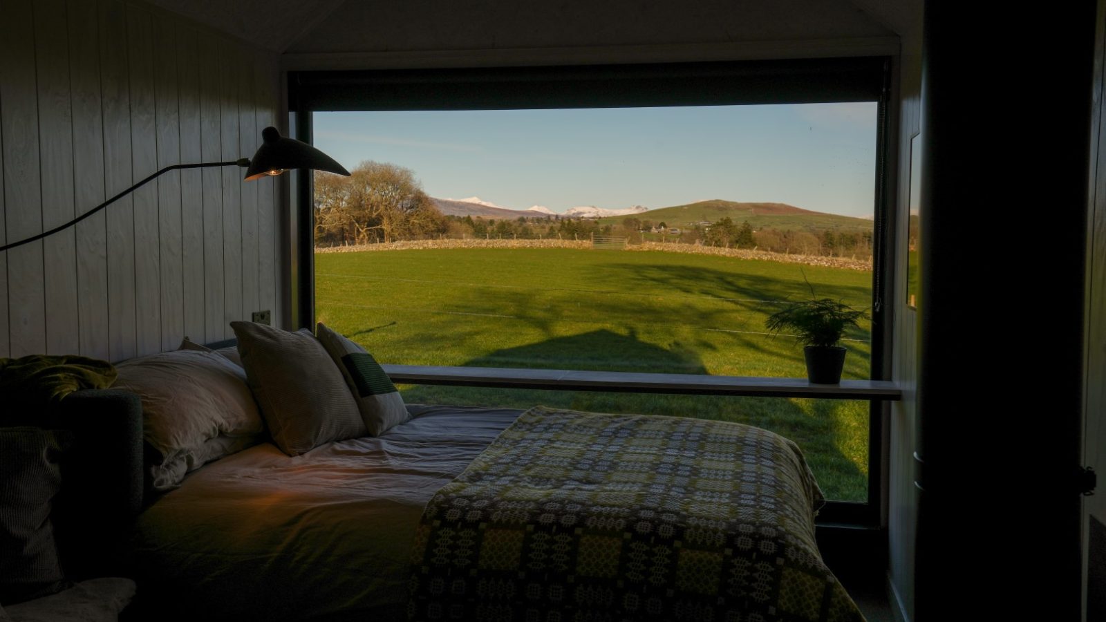 Bedroom with a large window view of a grassy field and distant hills; sunlight casts shadows across the bed and floor.