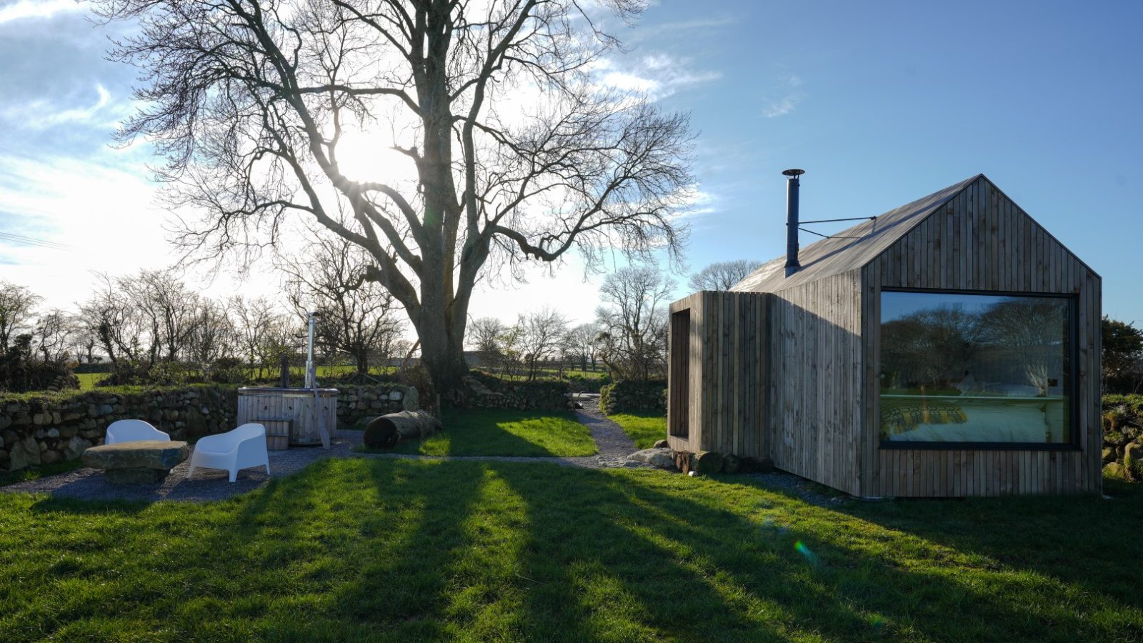 Small wooden cabin with a large window next to a leafless tree, set in a grassy field under a bright sun. Two chairs are nearby.