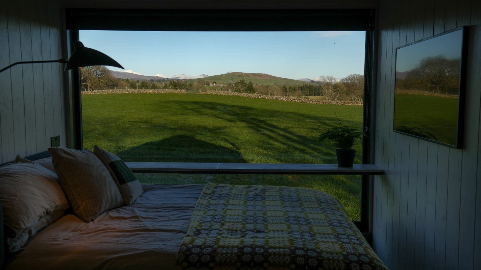 View from a bedroom through a large window overlooking a green field and distant hills under a clear blue sky.