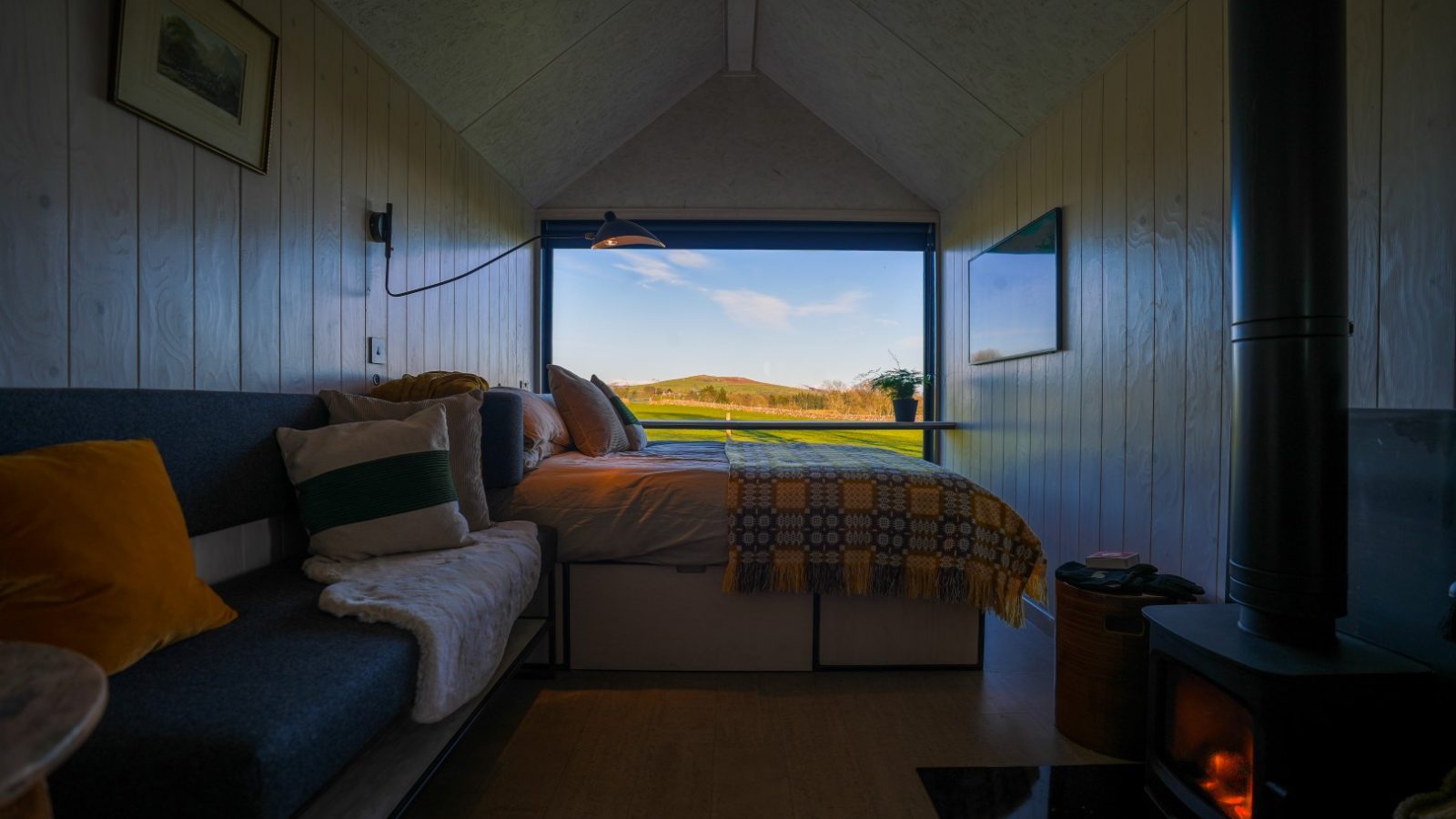 Cozy tiny home interior with a bed, sofa, and wood-burning stove. Large window shows a view of green fields and blue sky.