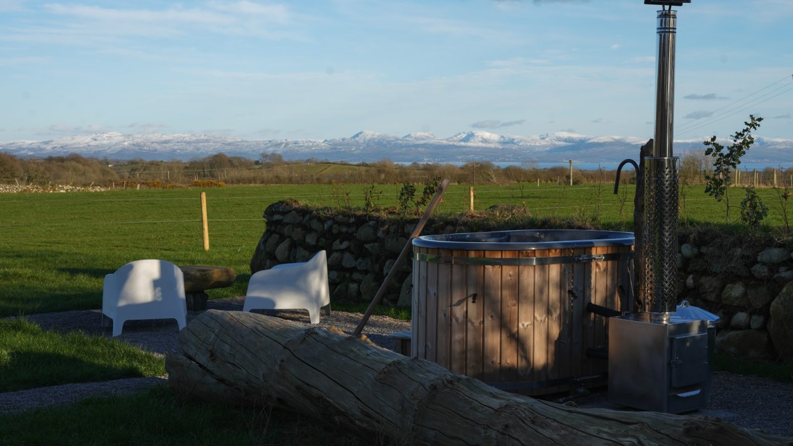 Outdoor hot tub and two white chairs on grass, with a log in the foreground and snow-capped mountains in the distance.