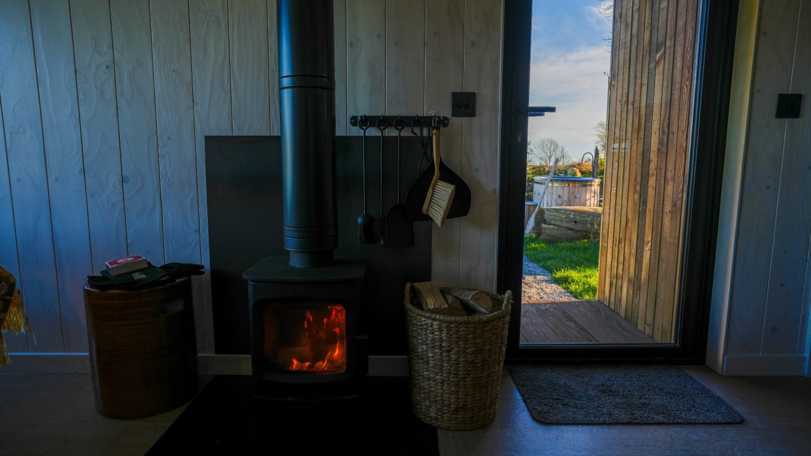 Wood stove with burning fire inside a room; basket of logs beside it. Open door reveals view of grass and outdoor scene.