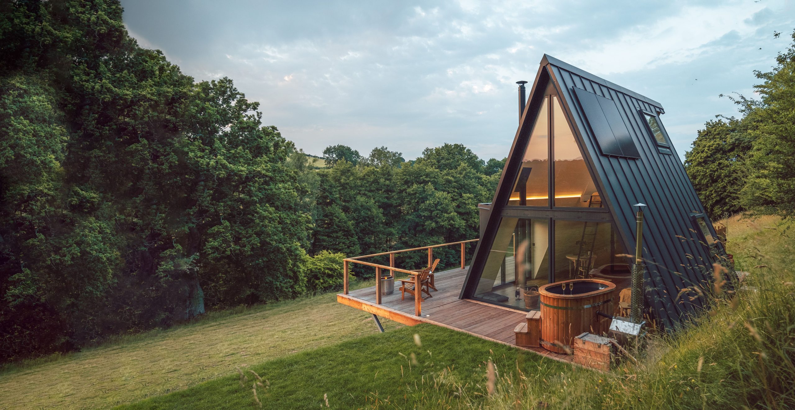 A-frame cabin with large windows and deck seating, hot tub for a digital detox stay amidst lush greenery.