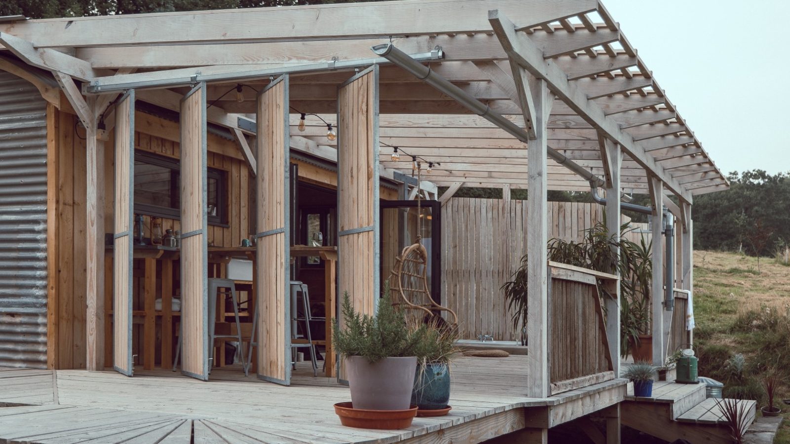 Wooden deck with potted plants opens to a Frith Meadow cabin, featuring a rustic metal and wood exterior.