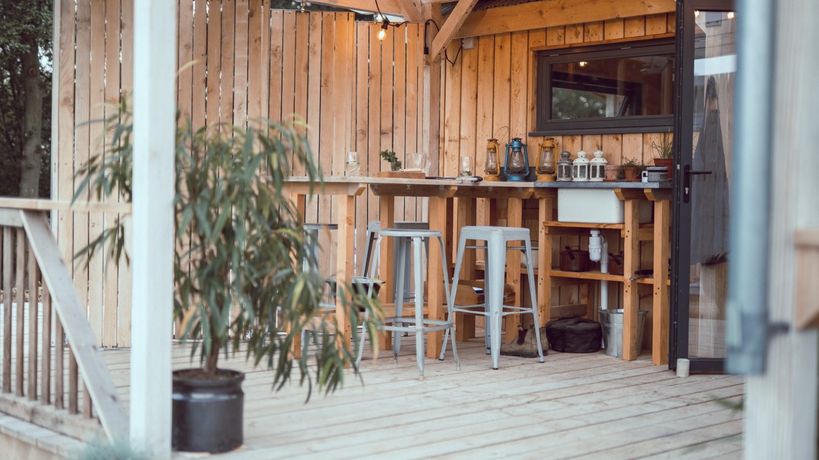 A wooden patio with bar stools, a counter, hanging lights, and a potted plant offers views of Carn through an open glass door.