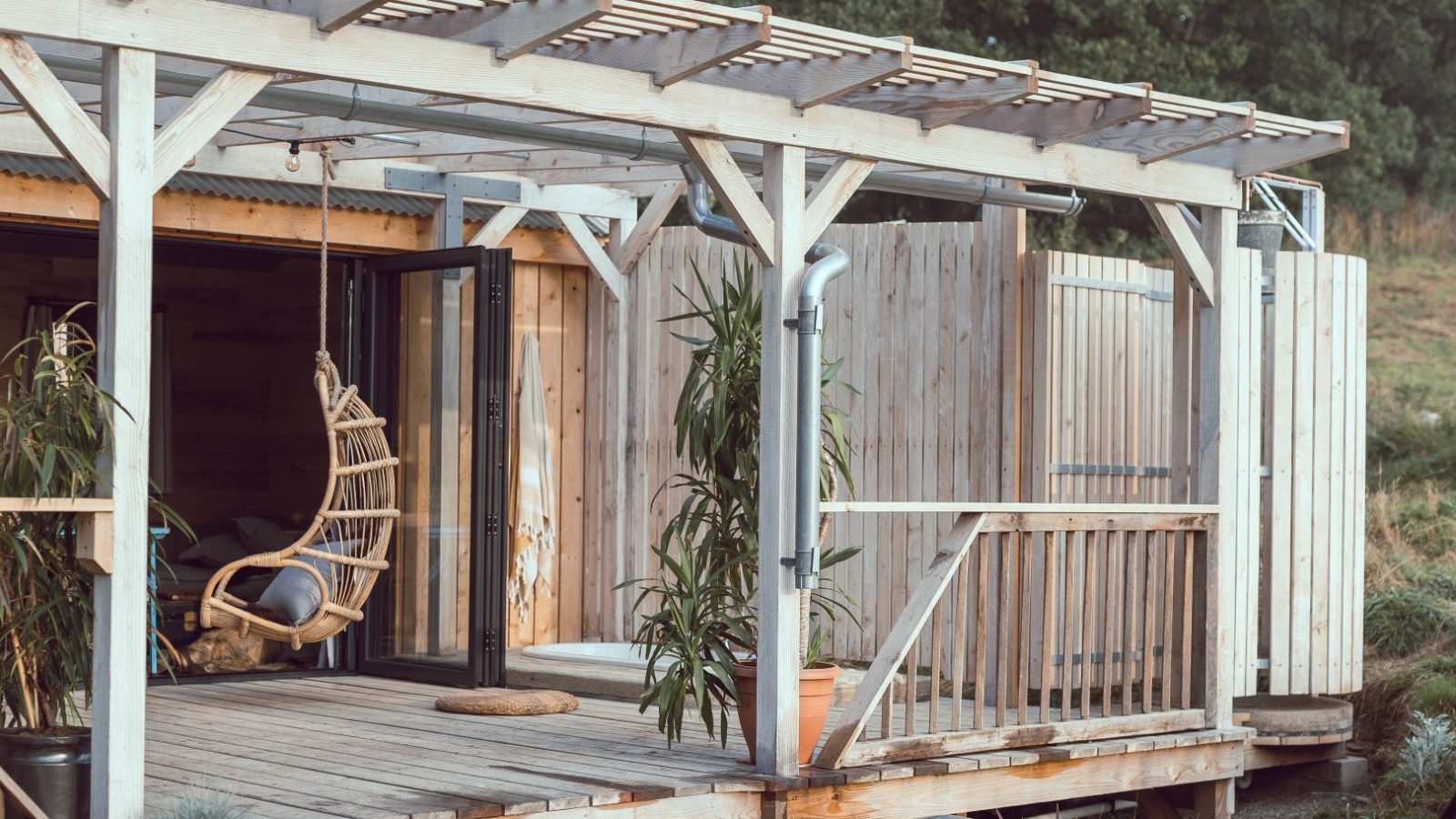 A wooden patio with a pergola, hanging chair, and plants opens to Frith Meadow's greenery through sliding doors.