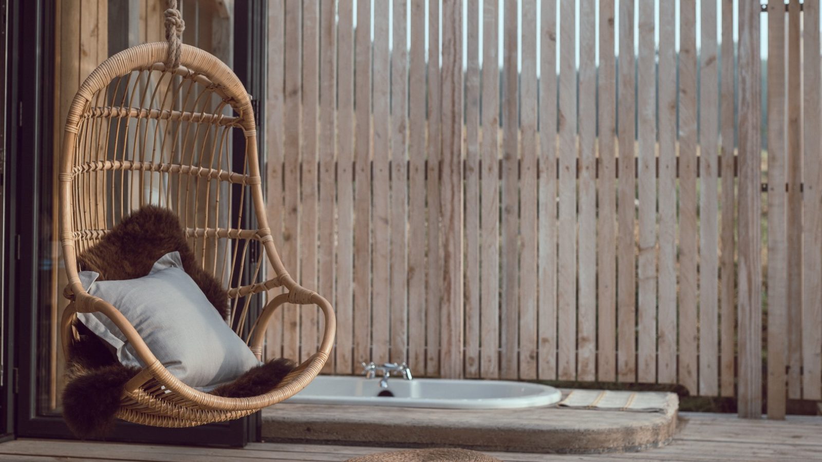 Hanging rattan chair with cushions on a wooden deck, overlooking Carn meadow, next to an outdoor bathtub and privacy screen.