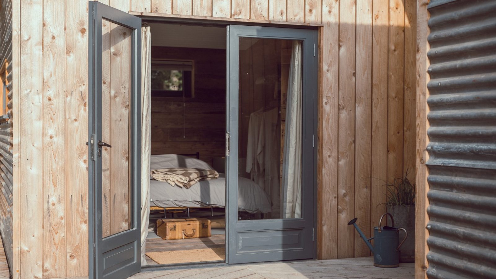 Open double doors lead to a Frith Meadow bedroom with a bed and suitcase. A watering can is on the wooden deck outside.