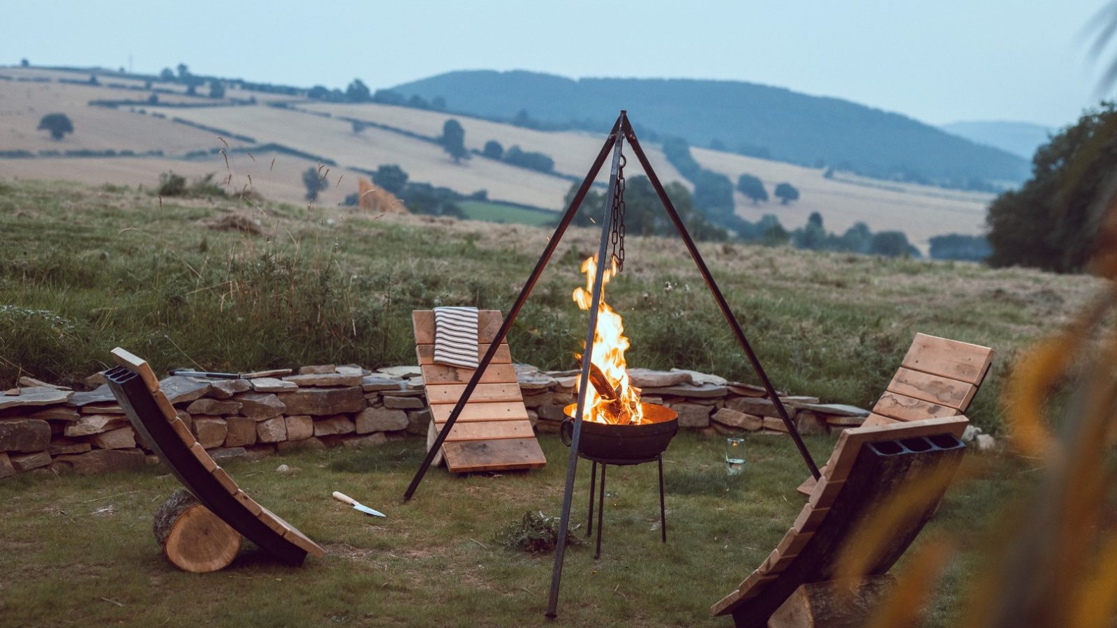 Three wooden lounge chairs around a tripod fire pit on Carn at Frith Meadow, with hills and a stone wall in the background.