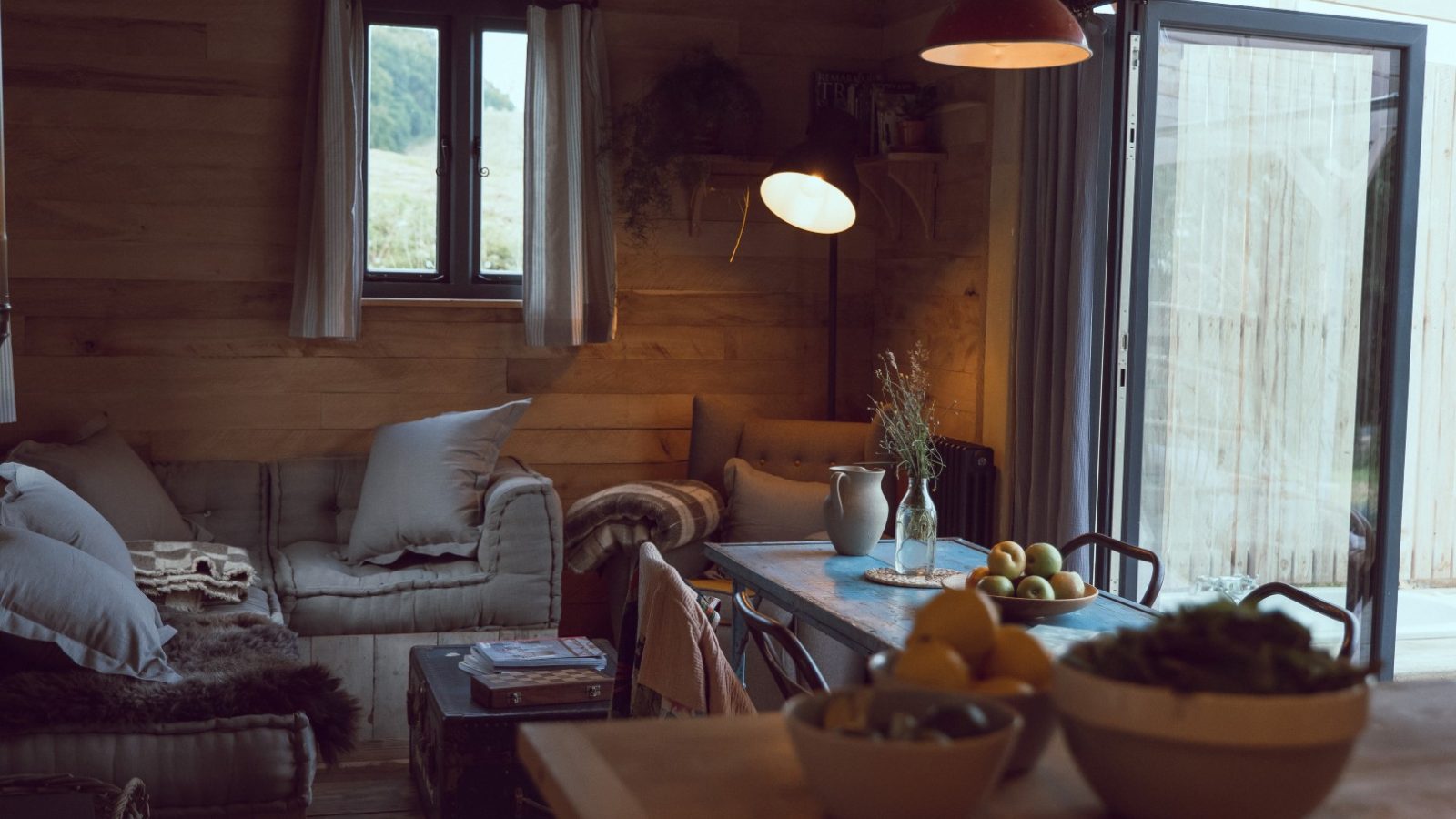 Cozy wooden interior in Frith Meadow with a sofa, fruit-laden table, and large windows. Soft lighting creates warmth.