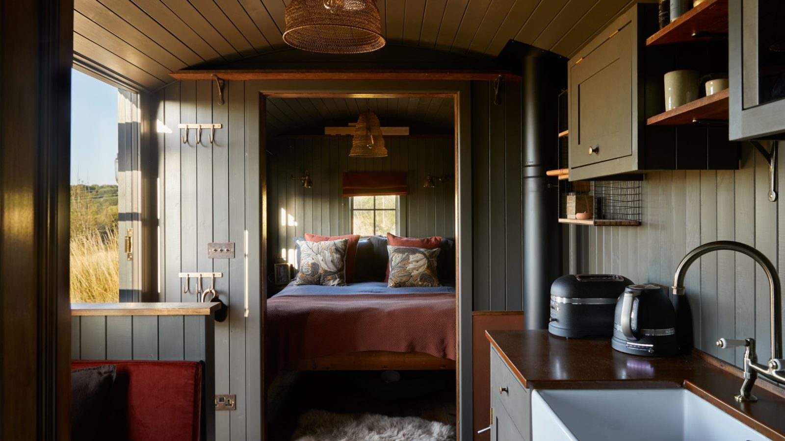 Cozy tiny house interior with a double bed, overhead light, kitchenette with sink and appliances, and wooden paneling.