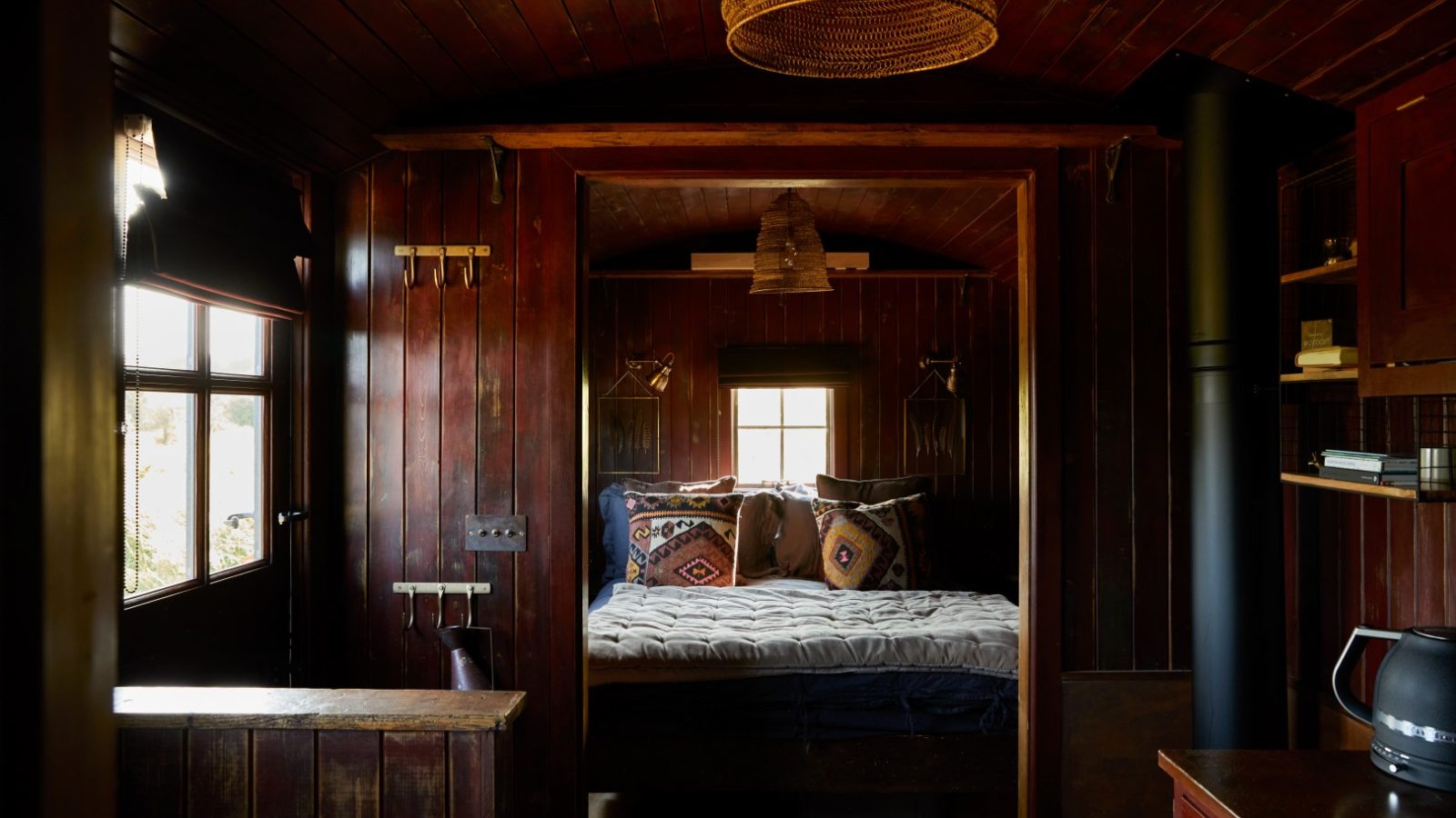 Cozy cabin interior with wooden walls, a bed with patterned cushions, a window, and ambient lighting from hanging fixtures.