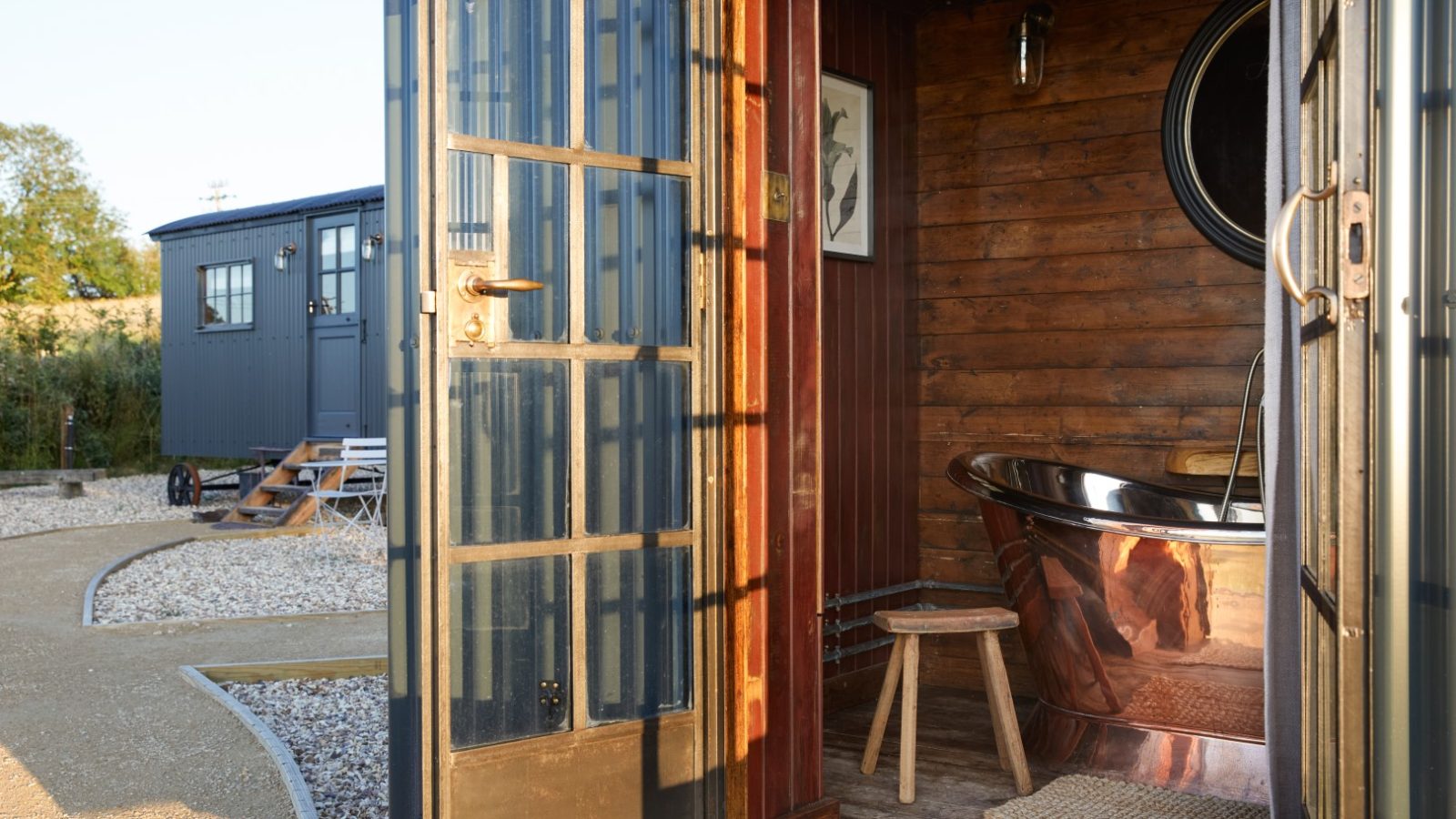 A rustic bathroom with a metal bathtub and wooden stool. The door is open, revealing a gravel path and additional structures outside.