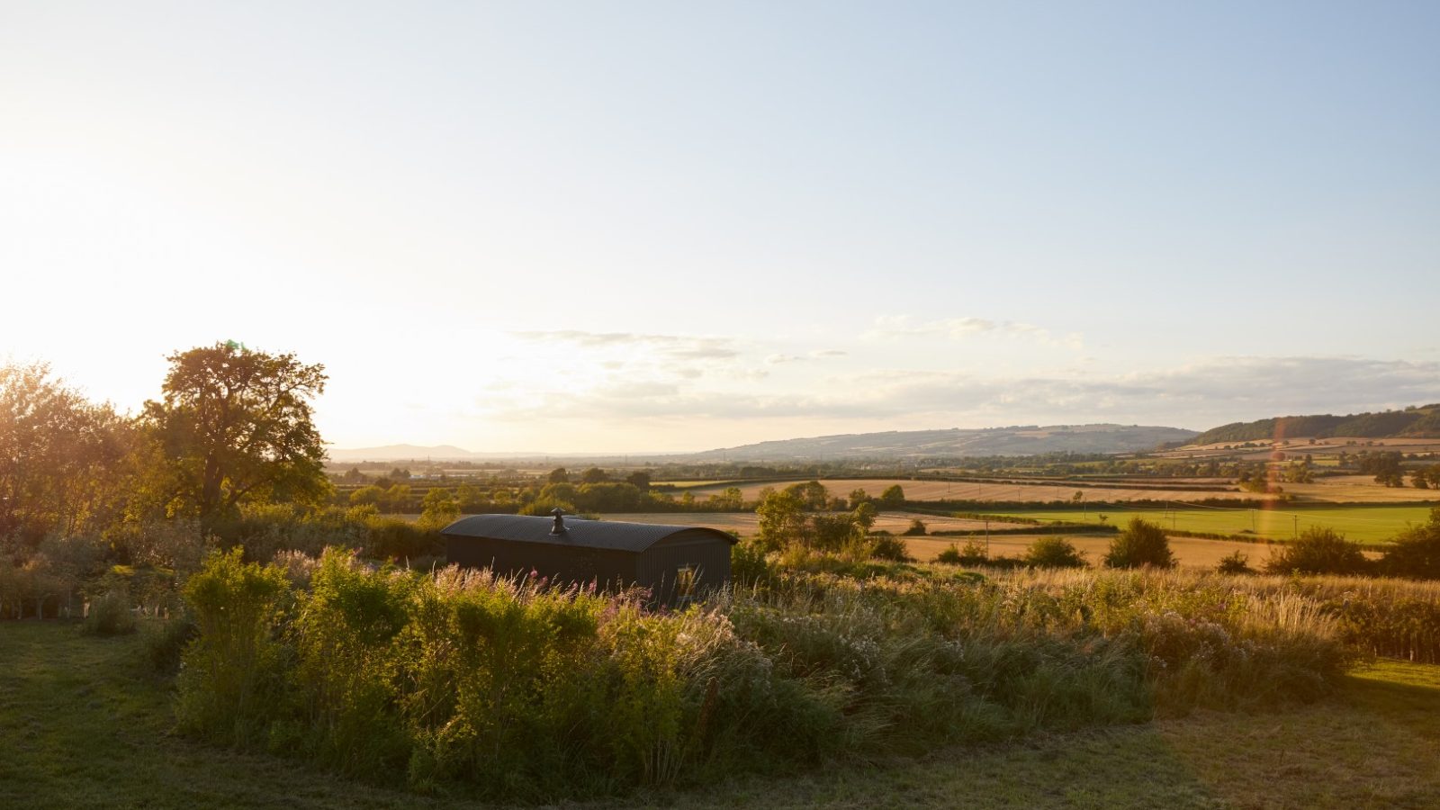 A scenic view of a rural landscape at sunset with a grassy field, a building, and distant hills under a clear sky.