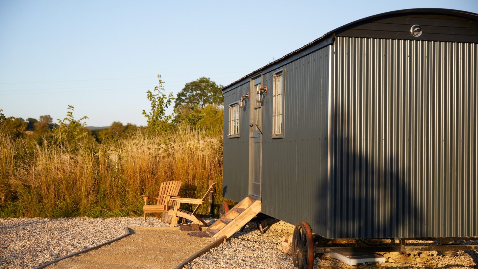 Small dark gray cabin on wheels with wooden chairs outside, set on a gravel path with shrubs and trees in the background.