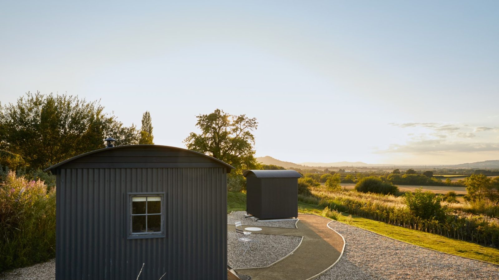 Two small dark-colored cabins on a gravel path overlook a scenic rural landscape at sunset, with trees and hills in the background.