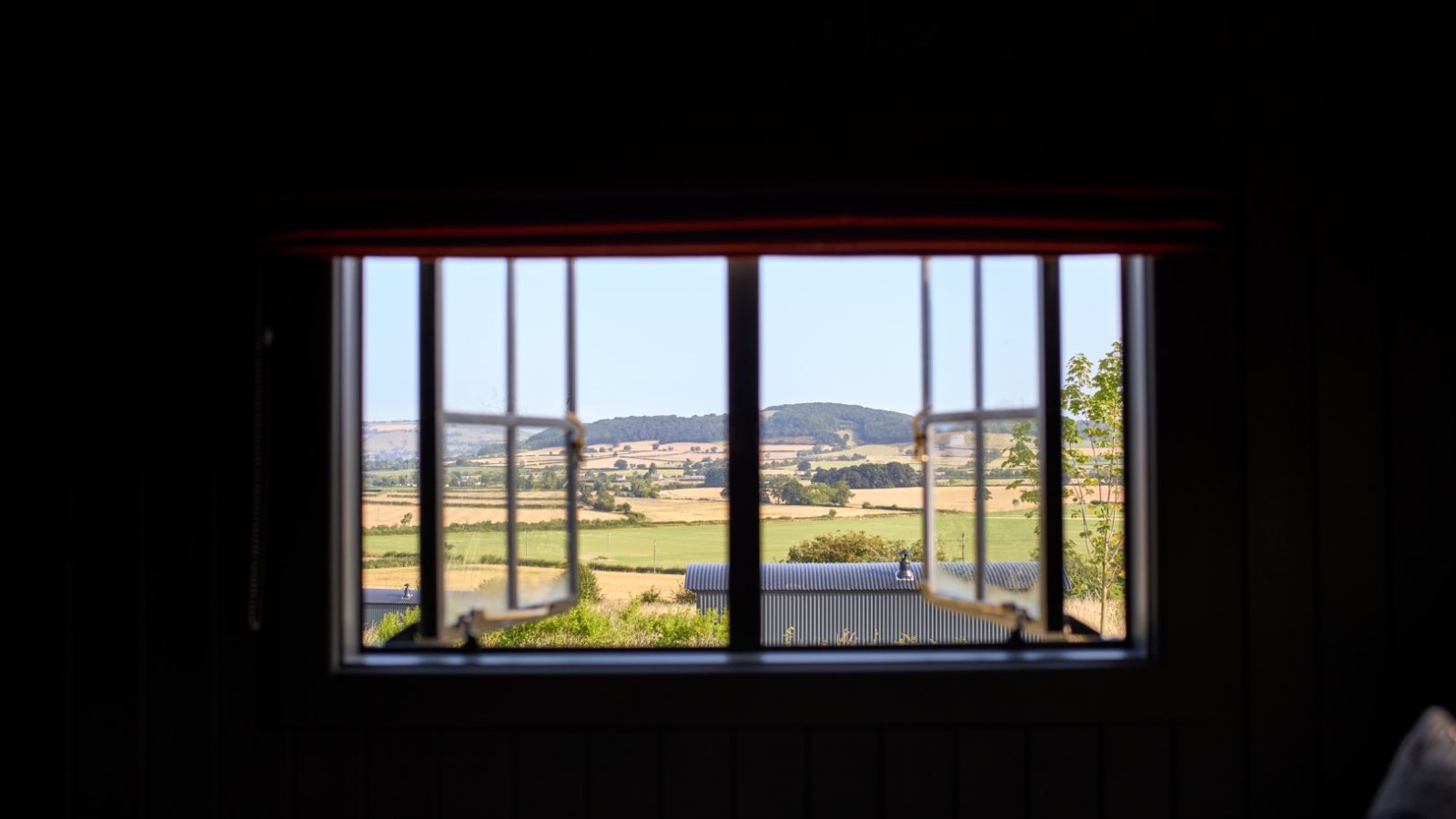 View of a rural landscape with fields and hills seen through an open window in a dark room.