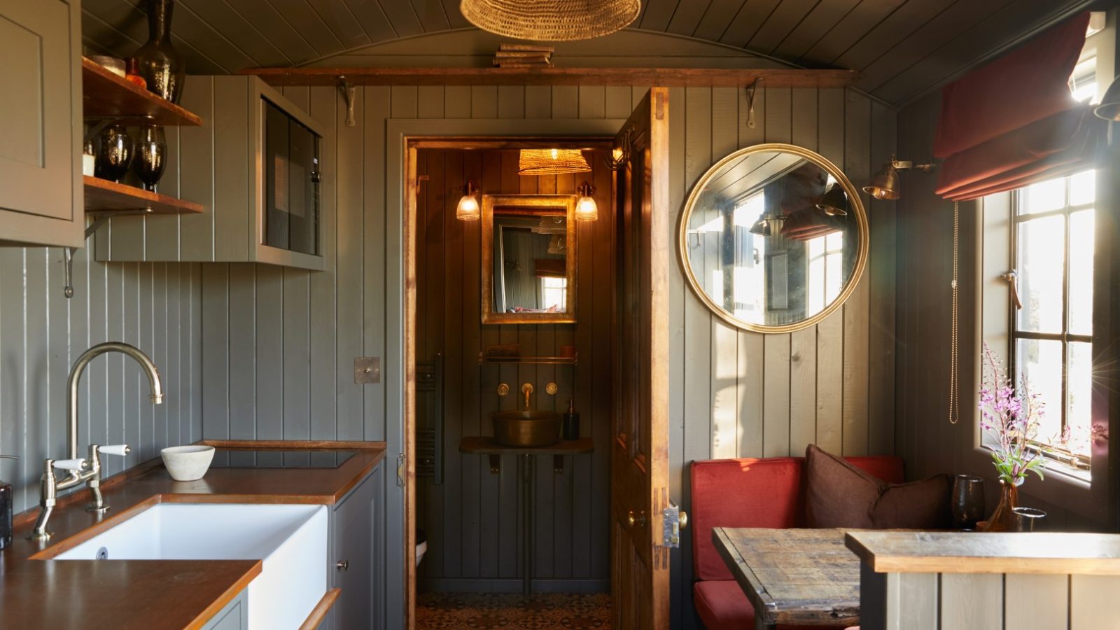 Cozy kitchen with wooden countertops, round mirror, and red cushioned bench. Door leads to a small, tiled bathroom.