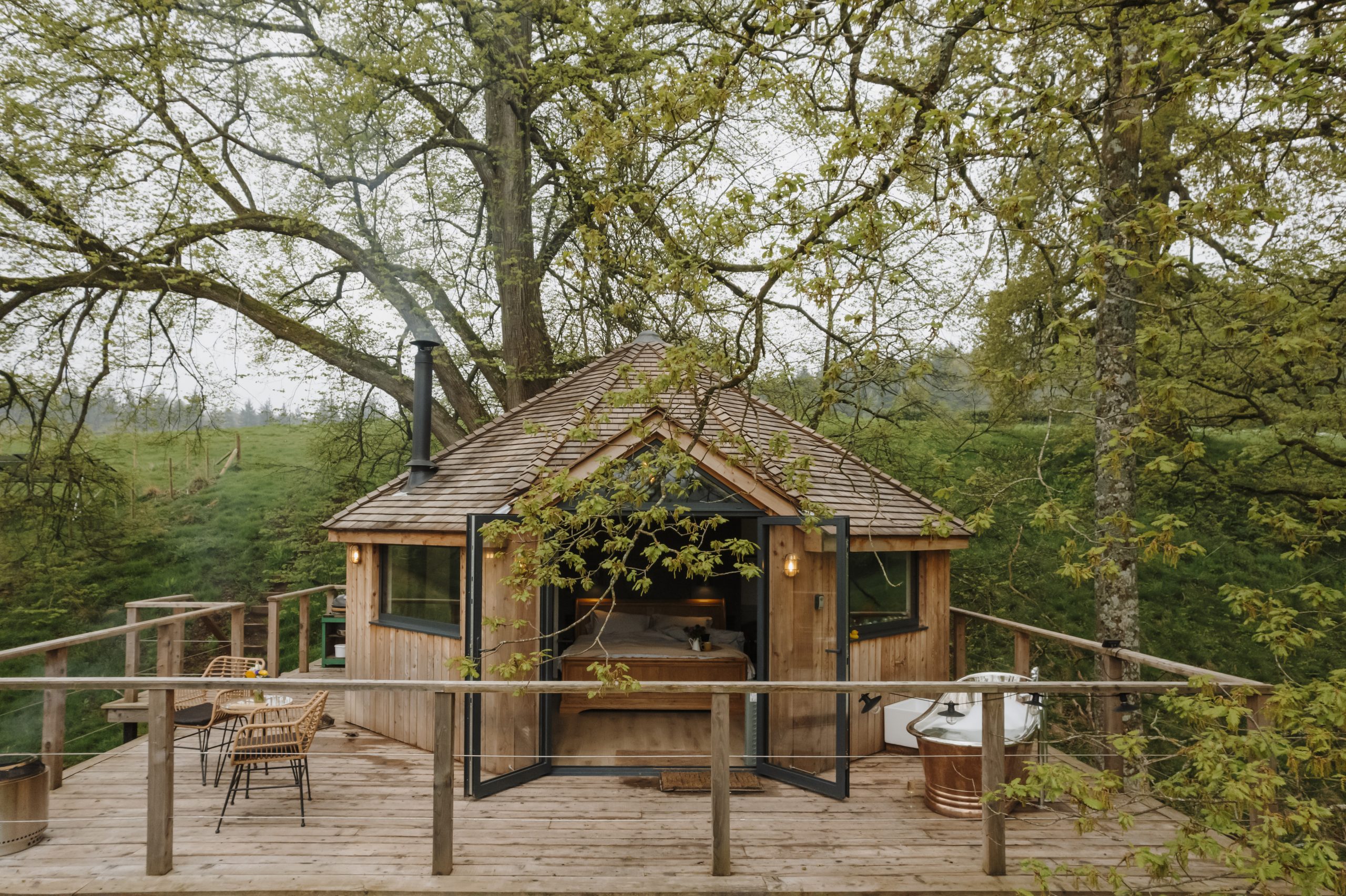 A romantic wooden cabin with open glass doors sits on a dog-friendly deck. There's outdoor seating and a bathtub under lush foliage.