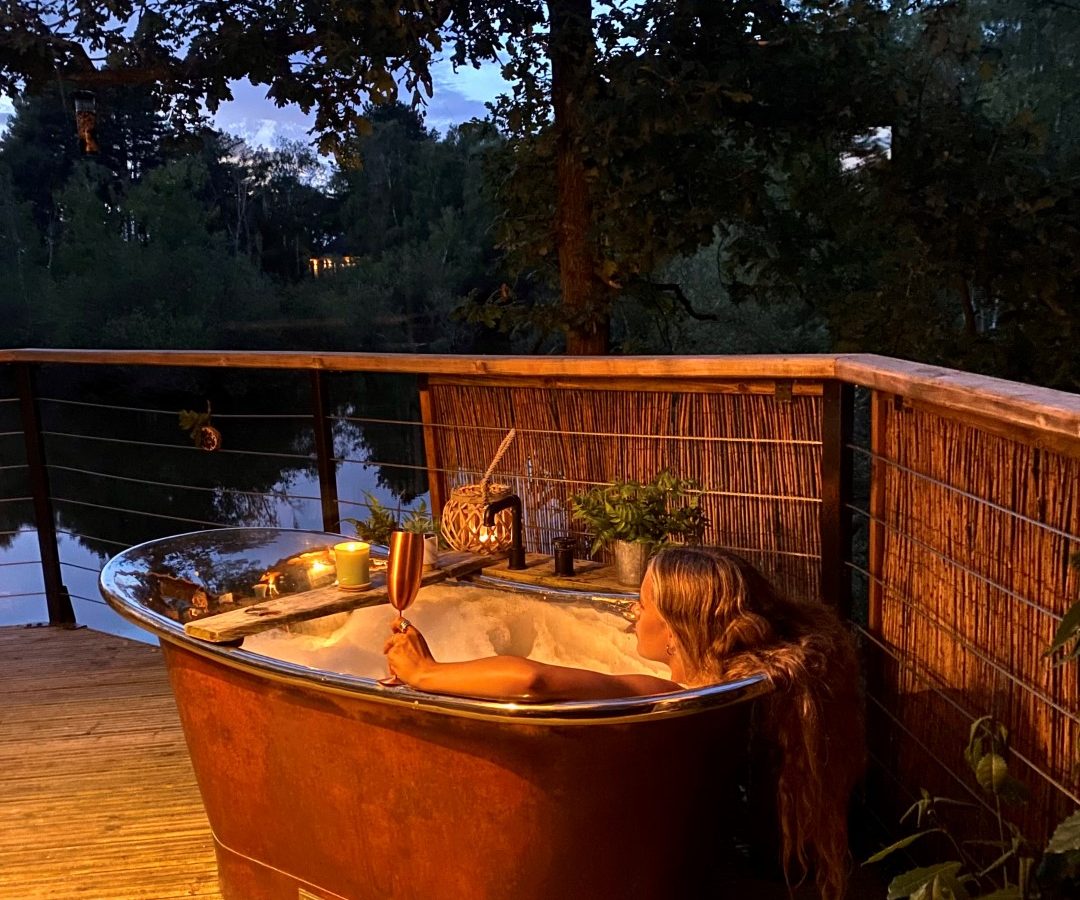 Relaxing in an outdoor copper bathtub at Treetop Hideaways, with a drink by dusk, surrounded by plants and lake views.