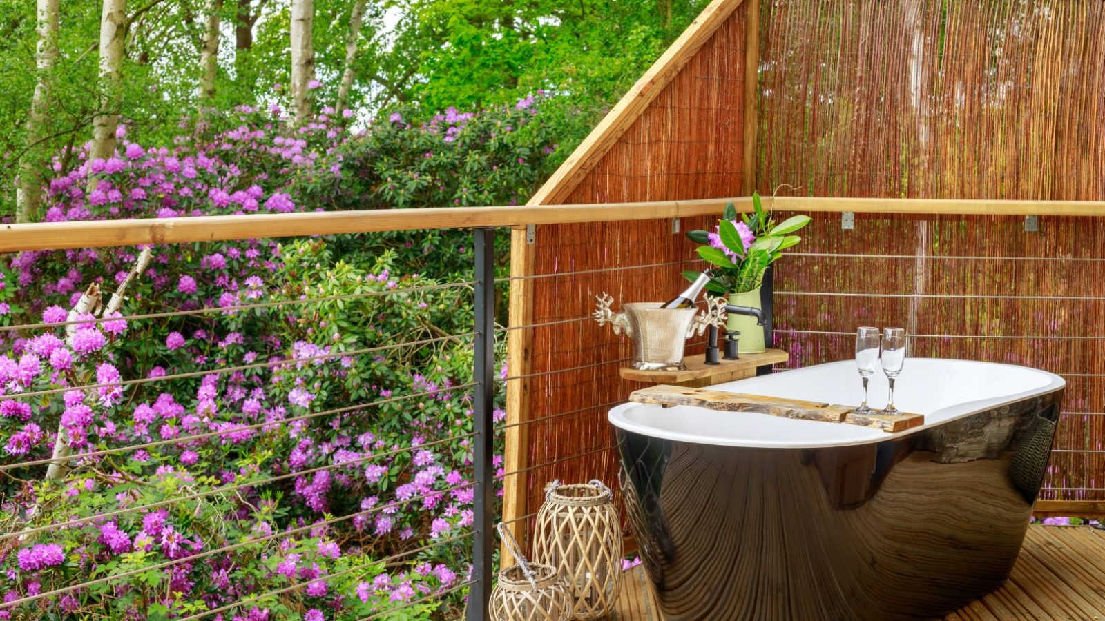 Treetop Hideaways: Outdoor bathtub on a wooden deck, purple flowers, two wine glasses, and lanterns create serene bliss.