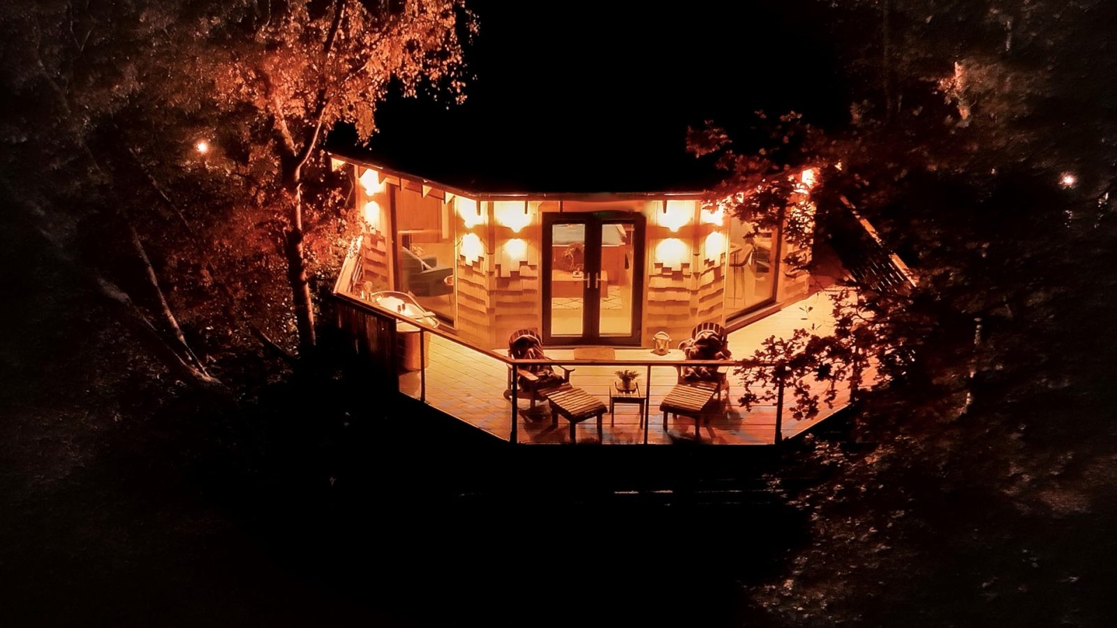 Aerial view of Treetop Hideaways' warmly lit hexagonal cabin at night, nestled among trees with deck chairs and tables.